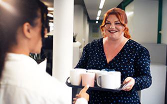 A woman carrying a tray with drinks and offering one to her colleague. A woman carrying a tray with drinks and offering one to her colleague.