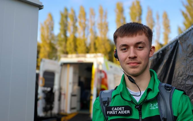 A Cadet First Aider wearing an earpiece in front of an ambulance. A Cadet First Aider wearing an earpiece in front of an ambulance.