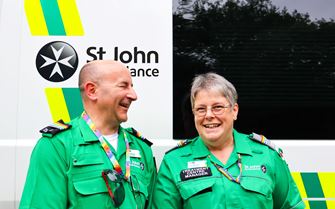 Two St John Ambulance volunteers in uniform smiling in front of an ambulance.