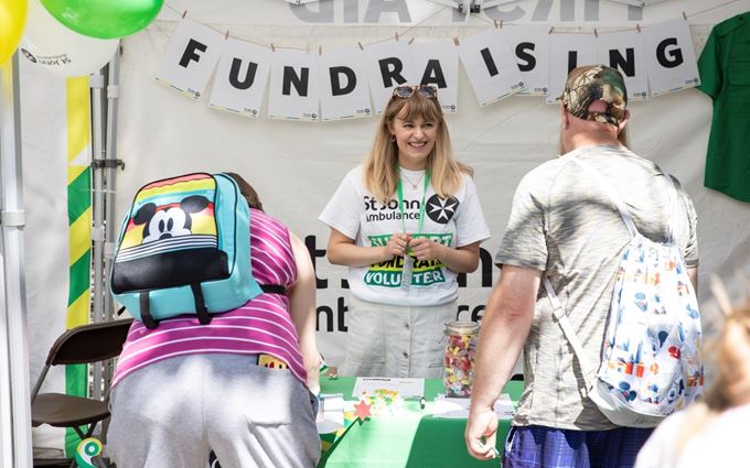 A volunteer standing at a table with a fundraising banner behind her and smiling at two members of the public.